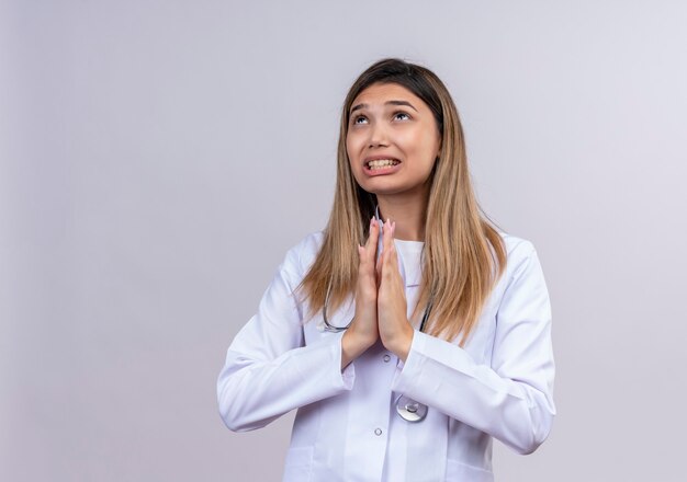 Joven hermosa mujer médico con bata blanca con estetoscopio pidiendo y orando con las manos junto con expresión de esperanza en la cara muy emocional y preocupada