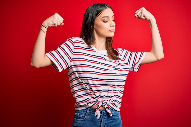 Foto gratuito joven hermosa mujer morena con una camiseta a rayas casual de pie sobre un fondo rojo mostrando los músculos de los brazos sonriendo orgulloso concepto de fitness