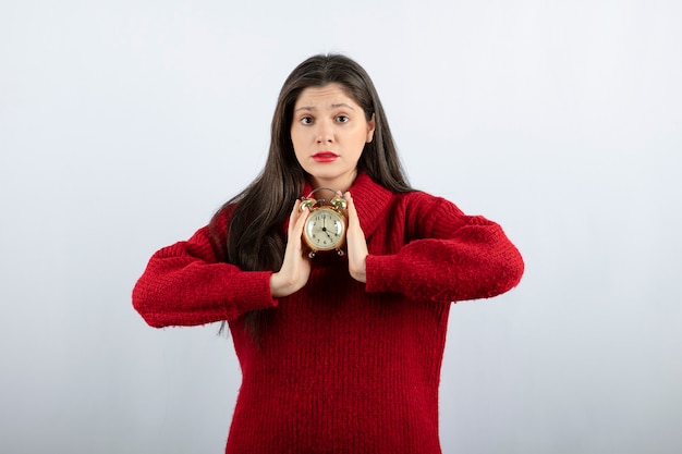 Joven hermosa mujer morena sosteniendo un reloj de alarma de pie sobre fondo blanco.