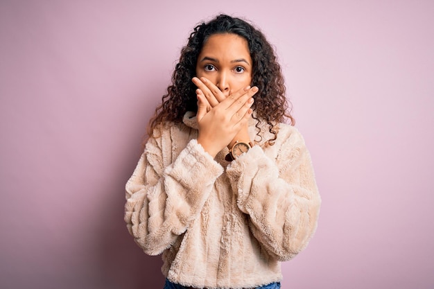 Foto gratuita joven hermosa mujer con el pelo rizado y un suéter informal de pie sobre fondo rosa sorprendido cubriéndose la boca con las manos por error concepto secreto