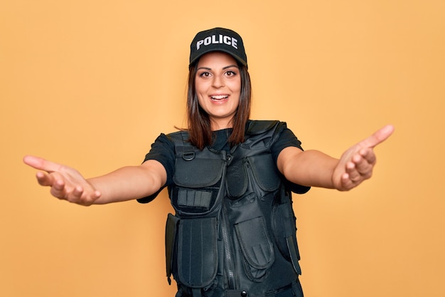 Foto gratuita joven y hermosa mujer policía morena con uniforme de policía a prueba de balas y gorra sonriendo alegre ofreciendo manos dando asistencia y aceptación