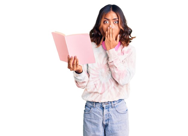 Joven hermosa mujer de raza mixta leyendo un libro con auriculares cubriendo la boca con la mano, conmocionada y asustada por el error. expresión sorprendida