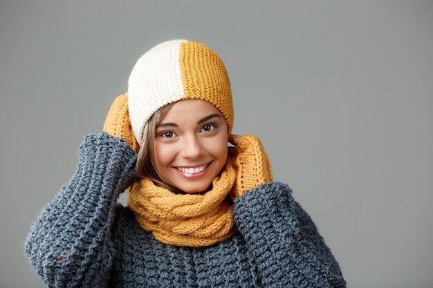 Joven hermosa mujer rubia en bufanda de punto sombrero suéter y mitones sonriendo en gris.