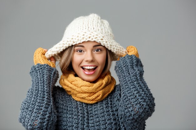 Joven hermosa mujer rubia en bufanda de punto sombrero suéter y mitones sonriendo en gris.