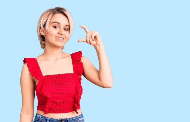 Joven hermosa mujer rubia con ropa informal sonriendo y confiada gesticulando con la mano haciendo un signo de tamaño pequeño con los dedos mirando y el concepto de medida de la cámara