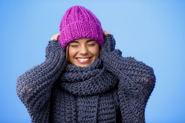 Joven hermosa mujer rubia en sombrero de punto y suéter sonriendo en azul.