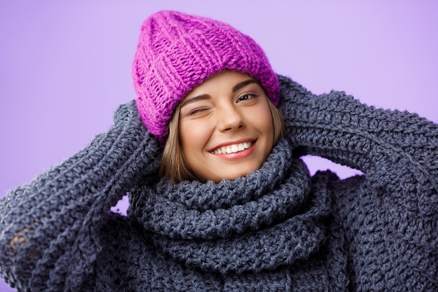 Joven hermosa mujer rubia en sombrero de punto y suéter sonriendo guiñando un ojo en violeta.