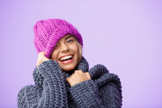 Joven hermosa mujer rubia en sombrero de punto y suéter sonriendo guiñando un ojo en violeta.