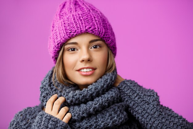 Joven hermosa mujer rubia con sombrero de punto y suéter sonriendo en violeta.