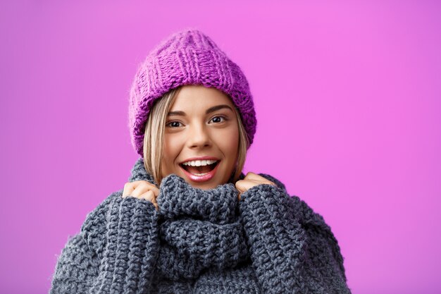 Joven hermosa mujer rubia con sombrero de punto y suéter sonriendo en violeta.
