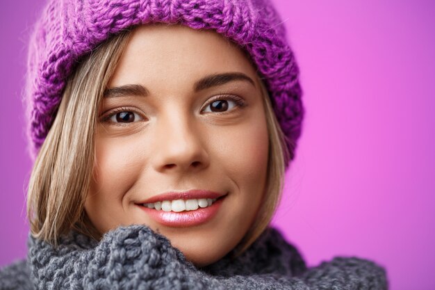 Joven hermosa mujer rubia con sombrero de punto y suéter sonriendo en violeta.