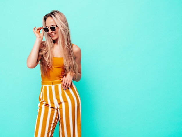 Foto gratuita joven hermosa mujer rubia sonriente en ropa amarilla de moda de verano mujer despreocupada sexy posando junto a la pared azul en el estudio modelo positivo divirtiéndose en el interior alegre y feliz en gafas de sol