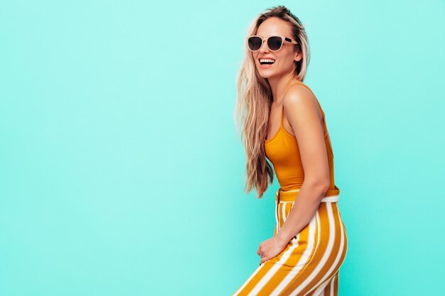 Joven hermosa mujer rubia sonriente en ropa amarilla de moda de verano Mujer despreocupada sexy posando junto a la pared azul en el estudio Modelo positivo divirtiéndose en el interior Alegre y feliz En gafas de sol