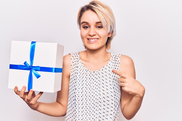 Joven hermosa mujer rubia sosteniendo regalo sonriendo feliz señalando con la mano y el dedo