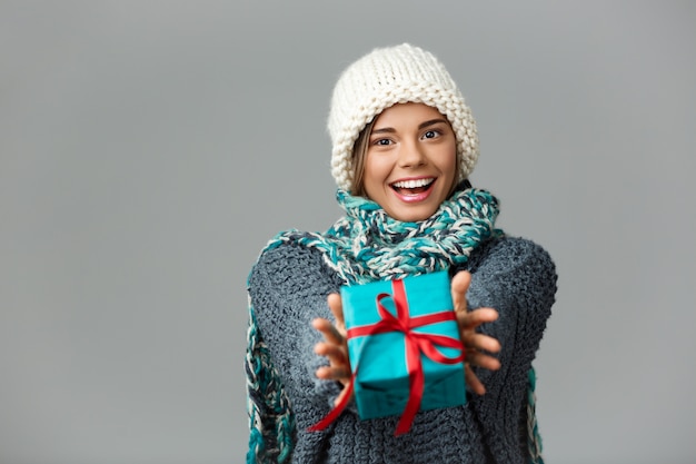 Joven hermosa mujer rubia en suéter de sombrero de punto y bufanda sonriendo dando caja de regalo en gris.