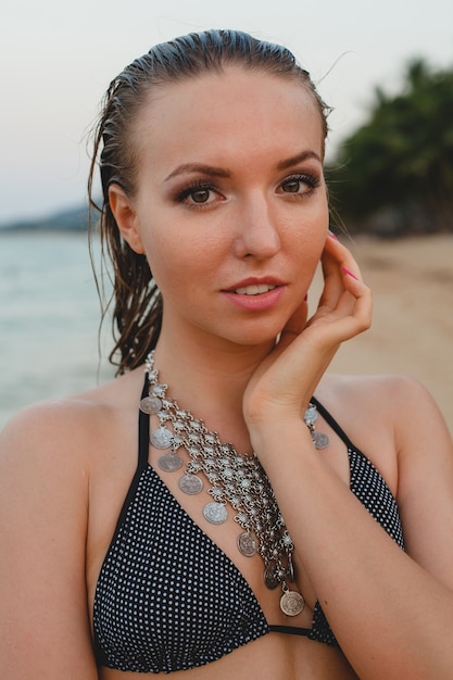 Joven hermosa mujer rubia tomando el sol en la playa de arena en traje de baño bikini, collar vintage