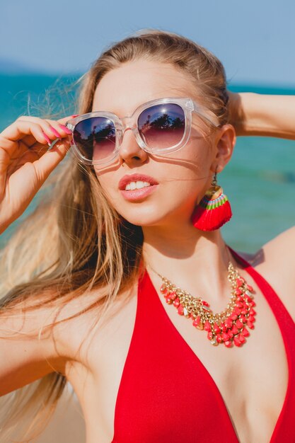 Joven hermosa mujer rubia tomando el sol en la playa en traje de baño rojo, gafas de sol