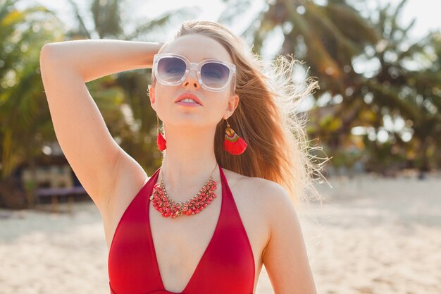 Joven hermosa mujer rubia tomando el sol en la playa en traje de baño rojo, gafas de sol