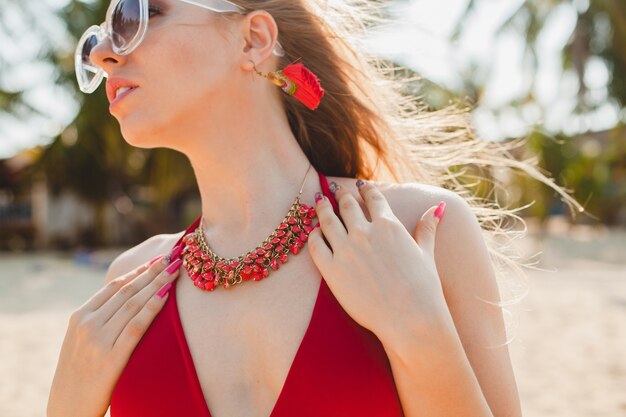 Joven hermosa mujer rubia tomando el sol en la playa en traje de baño rojo, gafas de sol