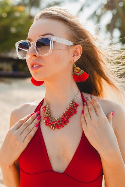 Joven hermosa mujer rubia tomando el sol en la playa en traje de baño rojo, gafas de sol