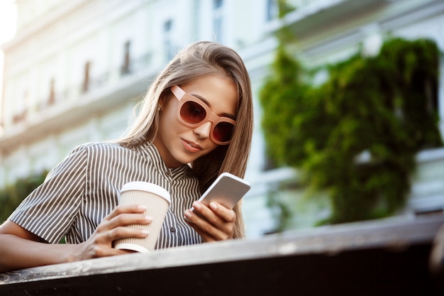 Joven hermosa mujer sentada en el banco, mirando el teléfono, sonriendo.