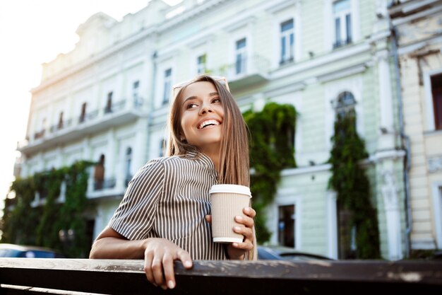 Joven hermosa mujer sentada en el banco, sosteniendo el café, sonriendo.