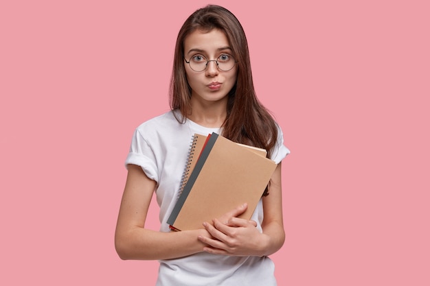 Joven hermosa mujer seria tiene cabello oscuro, lleva cuadernos de espiral, vestida con camiseta blanca, gafas, presiona los labios