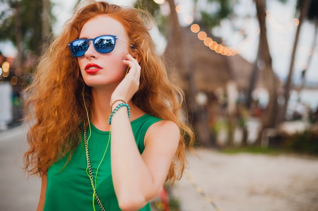 Joven hermosa mujer sexy, estilo hipster, cabello rojo, viajero, top verde, maleta naranja, vacaciones de verano, viajes, gafas de sol, escuchar música, auriculares, gafas de sol, viaje tropical