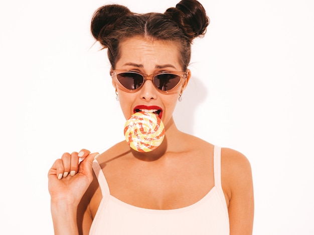 Joven hermosa mujer sexy hipster con labios rojos en gafas de sol. Chica de moda en ropa de baño de verano. Comer, morder paleta de caramelo