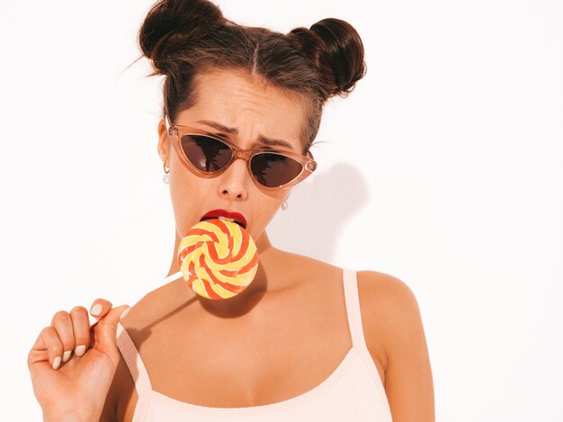 Joven hermosa mujer sexy hipster con labios rojos en gafas de sol. Chica de moda en ropa de baño de verano. Comer, morder paleta de caramelo