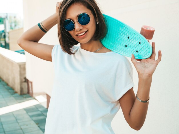 Joven hermosa mujer sexy hipster sonriente en gafas de sol. Chica de moda en camiseta y pantalones cortos de verano. Mujer positiva con patín azul centavo posando en la calle cerca de la pared blanca