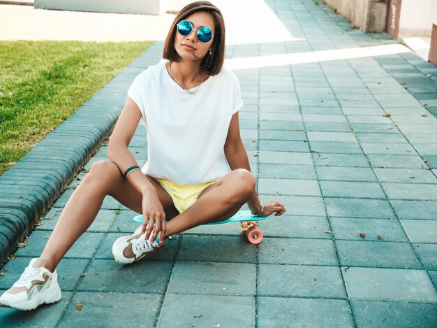 Joven hermosa mujer sexy hipster sonriente en gafas de sol. Chica de moda en camiseta y pantalones cortos de verano. Mujer positiva con patín azul centavo posando en el fondo de la calle