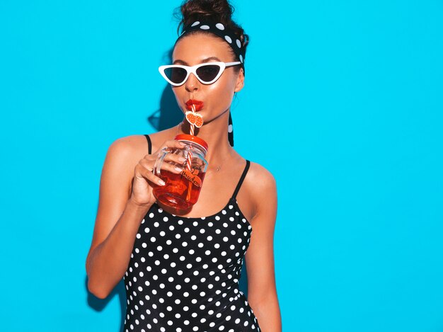 Joven hermosa mujer sexy hipster sonriente en gafas de sol. Chica en traje de baño de verano guisantes traje de baño. Colocando cerca de la pared azul, bebiendo cóctel fresco bebida lisa