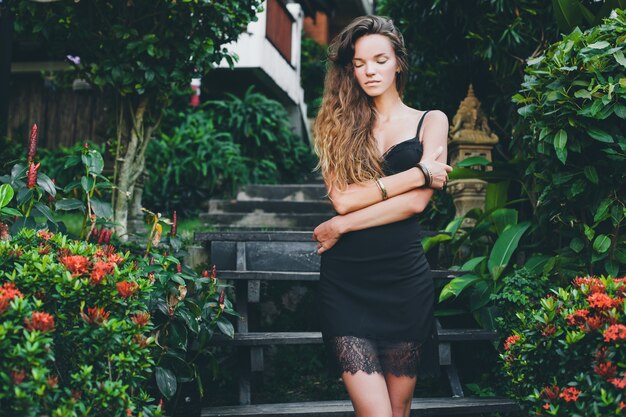 Joven hermosa mujer sexy en jardín tropical, vacaciones de verano en Tailandia, delgado cuerpo bronceado flaco, vestidito negro con encaje, aspecto natural, sensual, relajado,