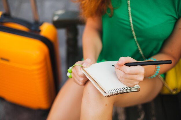 Joven hermosa mujer sexy, traje hipster, viajero, maleta naranja, haciendo notas en el libro de diario de viaje, vacaciones de verano, aventura, viaje, colorido, manos escribiendo, bolígrafo, detalles de cerca