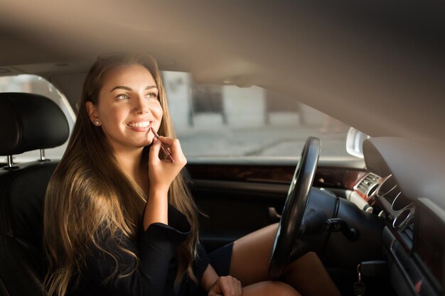 Joven hermosa mujer sonriente aplicando lápiz labial mientras está sentado en el auto detrás del volante