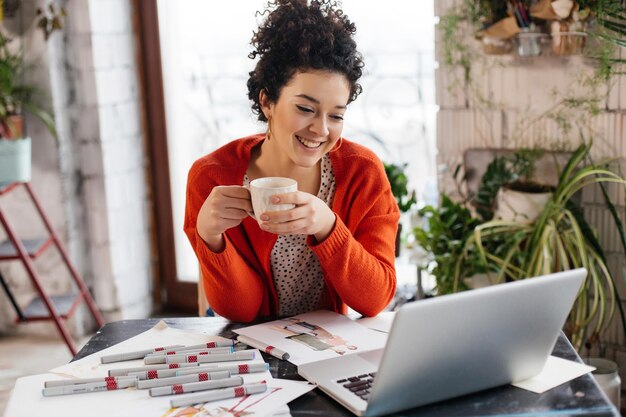 Joven hermosa mujer sonriente con cabello oscuro y rizado sentada en la mesa sosteniendo una taza en la mano felizmente trabajando en una laptop mientras pasa tiempo en un moderno y acogedor taller con grandes ventanas