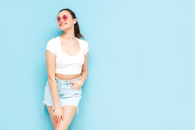 Joven hermosa mujer sonriente en falda de jeans de verano de moda y ropa superior Mujer despreocupada sexy posando junto a la pared azul en el estudio Modelo morena positiva divirtiéndoseAlegre y feliz