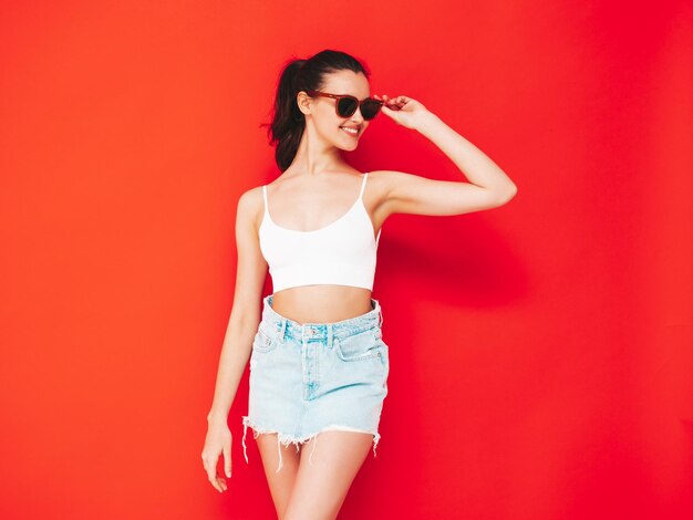 Joven hermosa mujer sonriente en falda y top de jeans de verano de moda Mujer despreocupada sexy posando junto a la pared roja en el estudio Modelo morena positiva divirtiéndose y volviéndose loca Alegre y feliz