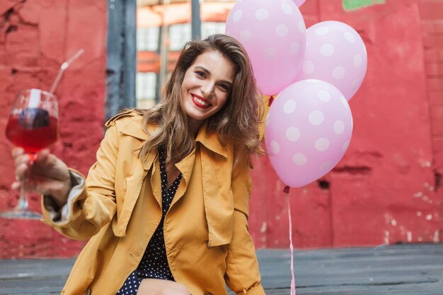 Joven hermosa mujer sonriente con gabardina naranja sosteniendo un cóctel en la mano mirando felizmente a la cámara con globos rosas cerca del antiguo y acogedor patio del café