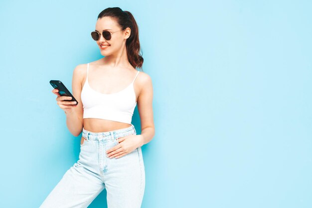 Joven hermosa mujer sonriente en jeans de moda de verano Mujer sexy despreocupada posando cerca de la pared azul en el estudio Modelo morena positiva mirando la pantalla del teléfono celular Sosteniendo el teléfono inteligente y usando aplicaciones