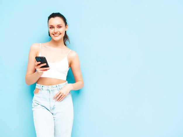 Joven hermosa mujer sonriente en jeans de moda de verano Mujer sexy despreocupada posando junto a la pared azul en el estudio Modelo morena positiva mirando la pantalla del teléfono celular Sosteniendo el teléfono inteligente y usando aplicaciones