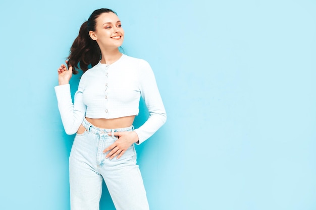 Joven hermosa mujer sonriente en jeans de moda de verano y ropa superior Sexy mujer despreocupada posando junto a la pared azul en el estudio Modelo morena positiva divirtiéndoseAlegre y feliz