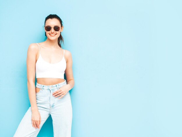 Joven hermosa mujer sonriente en jeans de moda de verano y ropa superior Sexy mujer despreocupada posando junto a la pared azul en el estudio Modelo morena positiva divirtiéndoseAlegre y feliz