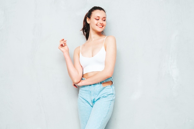 Joven hermosa mujer sonriente en jeans de moda de verano y top Sexy mujer despreocupada posando junto a la pared gris en el estudio Modelo morena positiva divirtiéndose y volviéndose loca Alegre y feliz