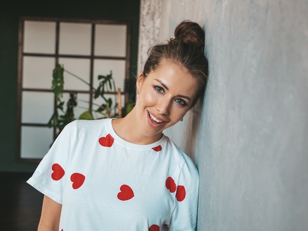 Joven hermosa mujer sonriente mirando a la cámara Chica de moda en camiseta blanca de verano casual Mujer divertida y positiva posando cerca de la pared gris en estudio