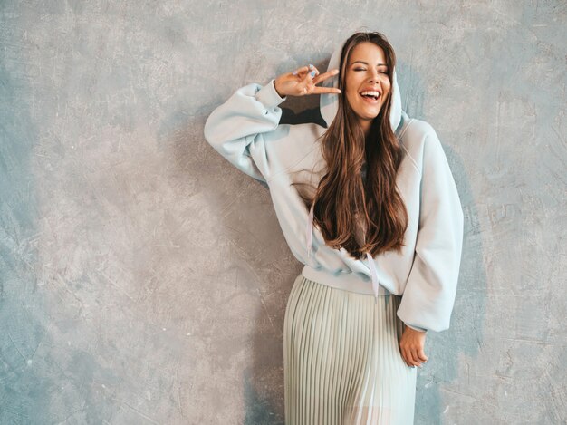 Joven hermosa mujer sonriente mirando. Chica de moda en ropa casual de verano con capucha y falda. Muestra un signo de paz.