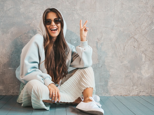 Joven hermosa mujer sonriente mirando. Chica de moda en ropa casual de verano con capucha y falda. Mujer divertida y positiva en gafas de sol sentados en el suelo y mostrando el signo de paz