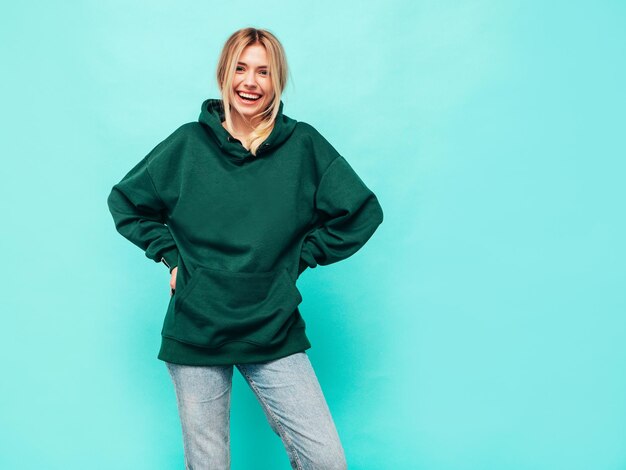 Joven hermosa mujer sonriente en moda sudadera con capucha verde de verano Sexy mujer despreocupada posando junto a la pared azul en el estudio Modelo rubio positivo divirtiéndose y volviéndose loco Alegre y feliz