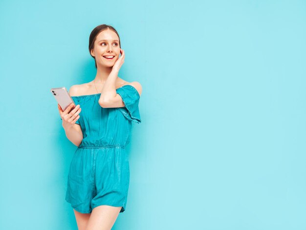 Joven hermosa mujer sonriente en monos azules de verano de moda Sexy mujer despreocupada posando cerca de la pared en el estudio Modelo morena positiva mirando la pantalla del teléfono celular Sosteniendo el teléfono inteligente y usando aplicaciones